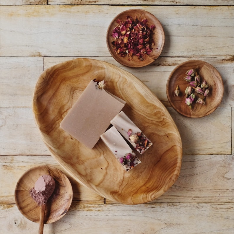 Blush colored Terra Rosa coconut milk soap bars are Naturally scented with a blend of Patchouli and geranium essential oils, laying in wooden bowl on a wooden table and surrounded by a small dish of rose clay and rose petals.