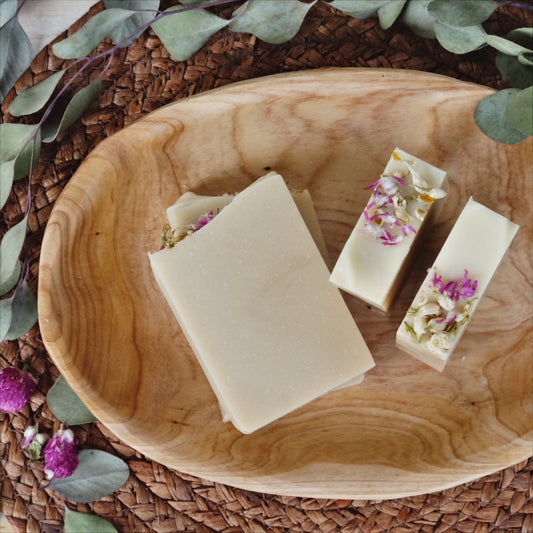 Creamy white bars of coconut milk soap scented with peppermint, eucalyptus and cedarwood atlas essential oils. Naturally colored with colloidal oatmeal, kaolin clay and plant oils, laying in a wooden bowl surrounded by eucalyptus leaves and flowers