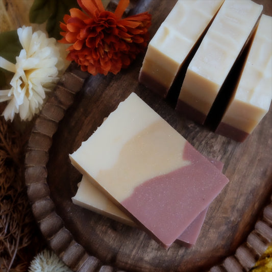 Handcrafted rectangular bars of soap with a cream and dusty rose swirl design, arranged on a dark wooden tray with dried flowers.