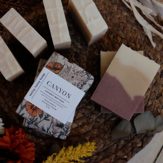 soap bar wrapped in earthy floral tissue paper with white band label next to unwrapped layered soap bars on brown mat and dried florals
