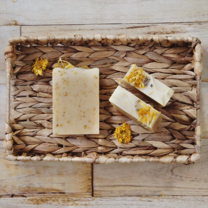 Golden Glow coconut milk soap bars laying in a woven basket on a wooden table. Scented with Lemongrass and lavender and naturally colored with calendula petals, colloidal oatmeal and kaolin clay. 
