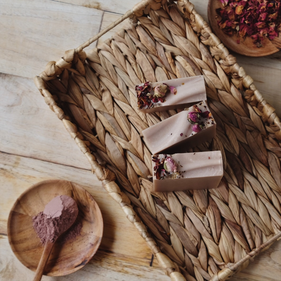 Blush colored Terra Rosa coconut milk soap bars are Naturally scented with a  blend of Patchouli and geranium essential oils, standing in woven basket and surrounded by rose clay and rose petals.