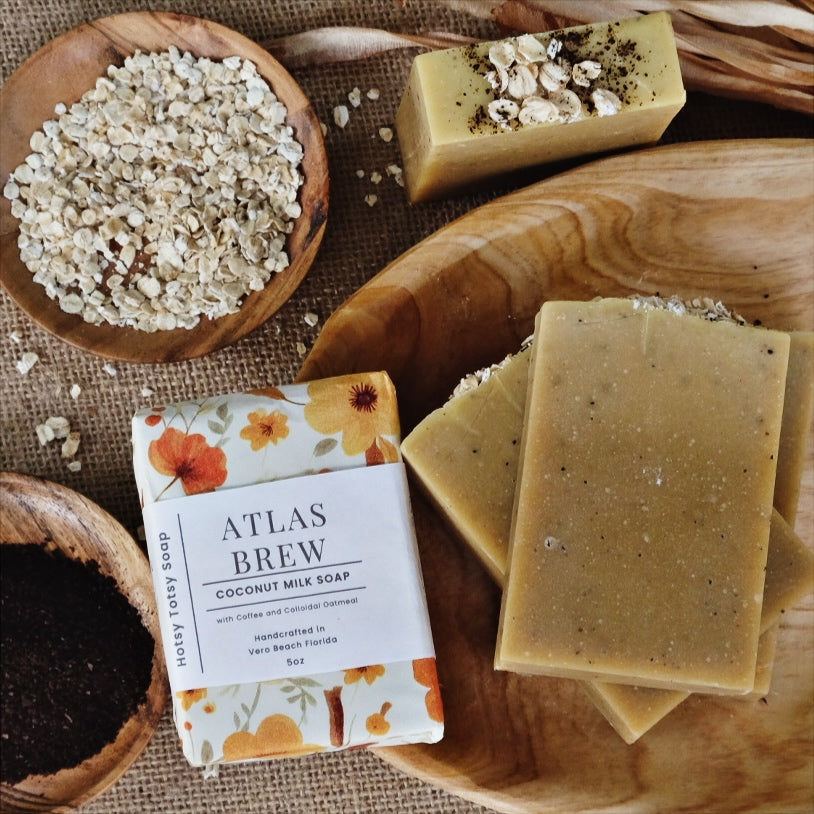Tan and speckled Atlas Brew Coconut milk soap bars  laying in a bowl surrounded by oats and coffee beans. Naturally scented with Cedarwood Atlas and orange essential oils. Each bar is wrapped in a  floral paper and labeled with a cigar band. 