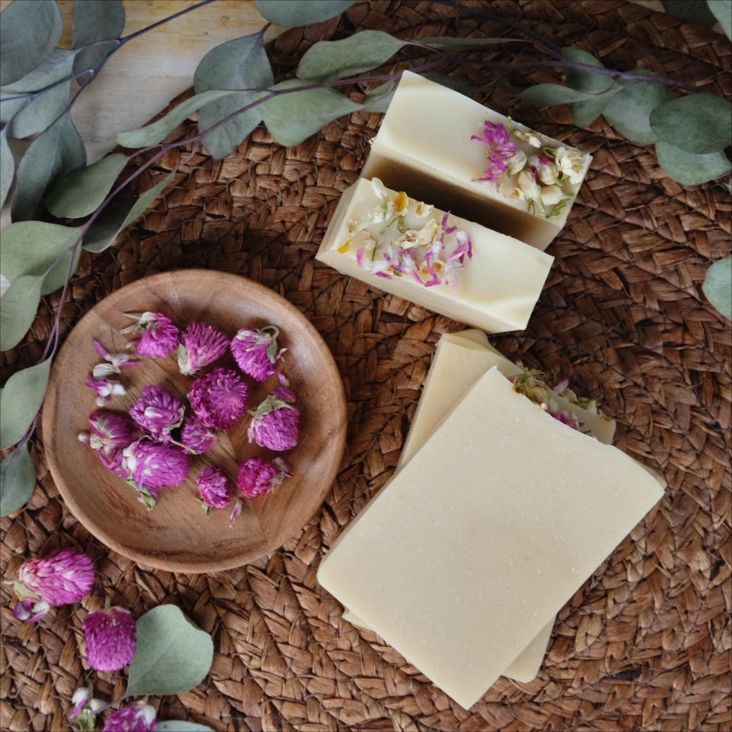 Creamy white bars of coconut milk soap scented with  peppermint, eucalyptus and cedarwood atlas essential oils, laying in a wooden bowl surrounded by eucalyptus leaves and flowers