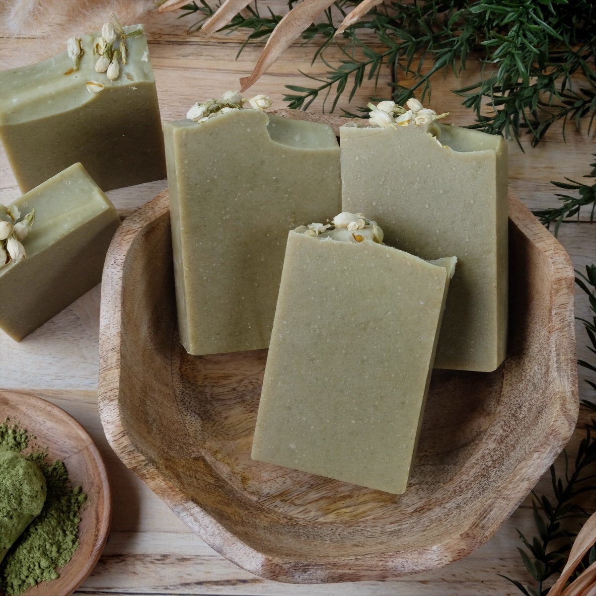 Green Meadow Mint coconut milk soap bars are naturally colored with organic spinach powder and Moringa leaf. Scented with a blend of peppermint and spearmint essential oils and decorated with dried jasmine flowers. Bar soaps are arranged in a wooden bowl with greenery surrounding.