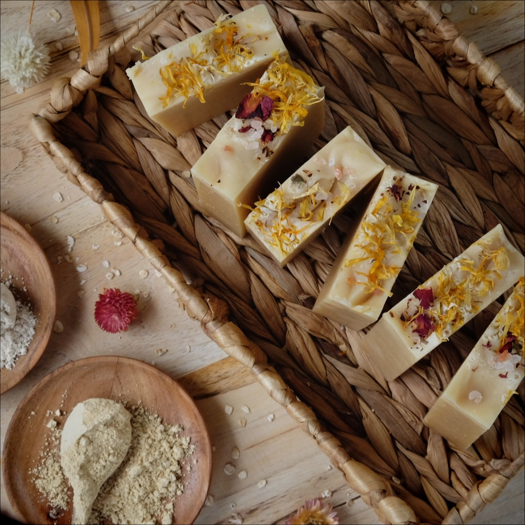Light yellow coconut milk soap bars scented with lavender, litsea cubeba and geranium essential oils and Naturally colored with yellow clay, colloidal oatmeal and kaolin clay standing in a woven basket surrounded by botanicals.