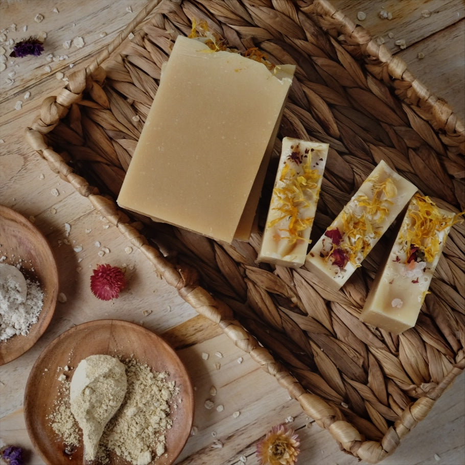 Light yellow coconut milk soap bars scented with lavender, litsea cubeba and geranium essential oils and Naturally colored with yellow clay, colloidal oatmeal and kaolin clay laying in a woven basket surrounded by botanicals.