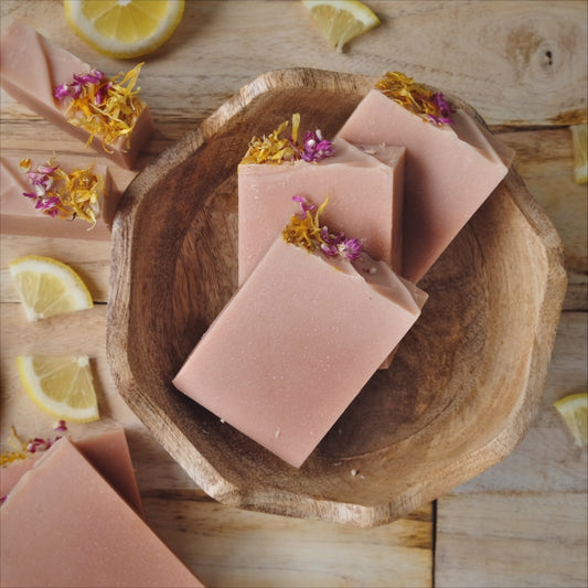 coconut milk soap bars stacked in a wooden bowel, naturally colored with French Pink clay. Scented with a blend of Bergamot, Litsea cubeba, and frankincense essential oils and decorated with dried flowers.
