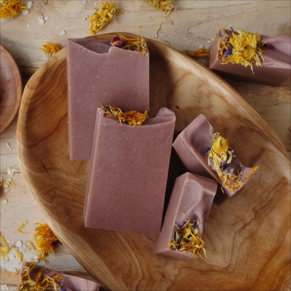 All natural soap bars colored with Moroccan red clay and scented with clove, cedarwood atlas, and patchouli essential oils and decorated with dried wild flowers and placed in a wooden bowl.