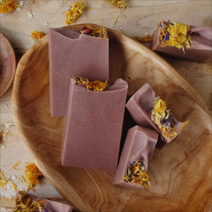 All natural soap bars colored with Moroccan red clay and scented with clove, cedarwood atlas, and patchouli essential oils and decorated with dried wild flowers and placed in a wooden bowl.