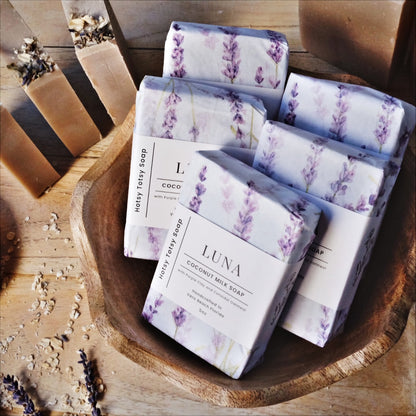 Luna coconut milk soap naturally colored with purple clay and scented with lavender essential oil. Soap bars are wrapped in white paper with purple lavender flowers.