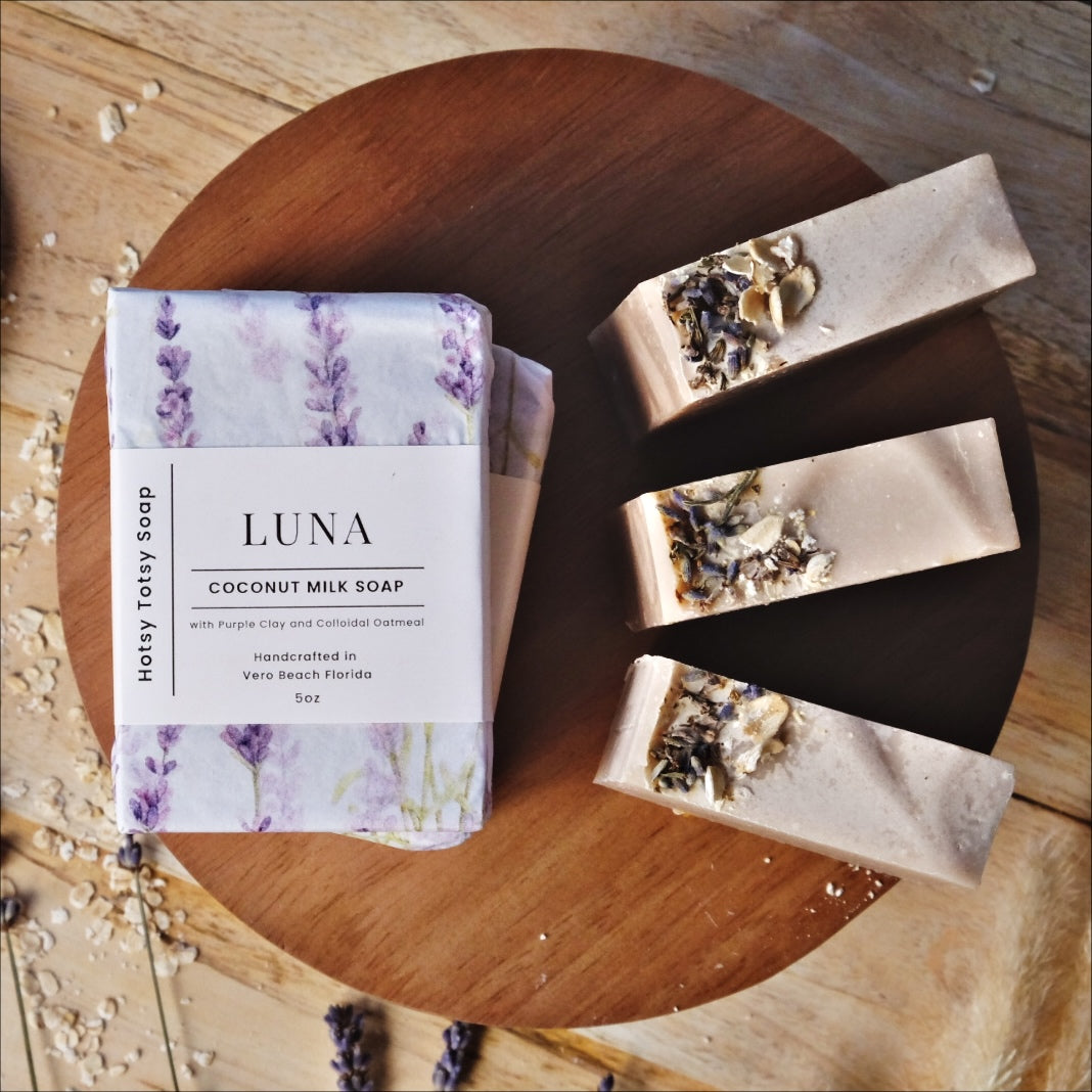 Luna coconut milk soap naturally colored with purple clay and scented with lavender essential oil. Soap bars are wrapped in white paper with purple lavender flowers.